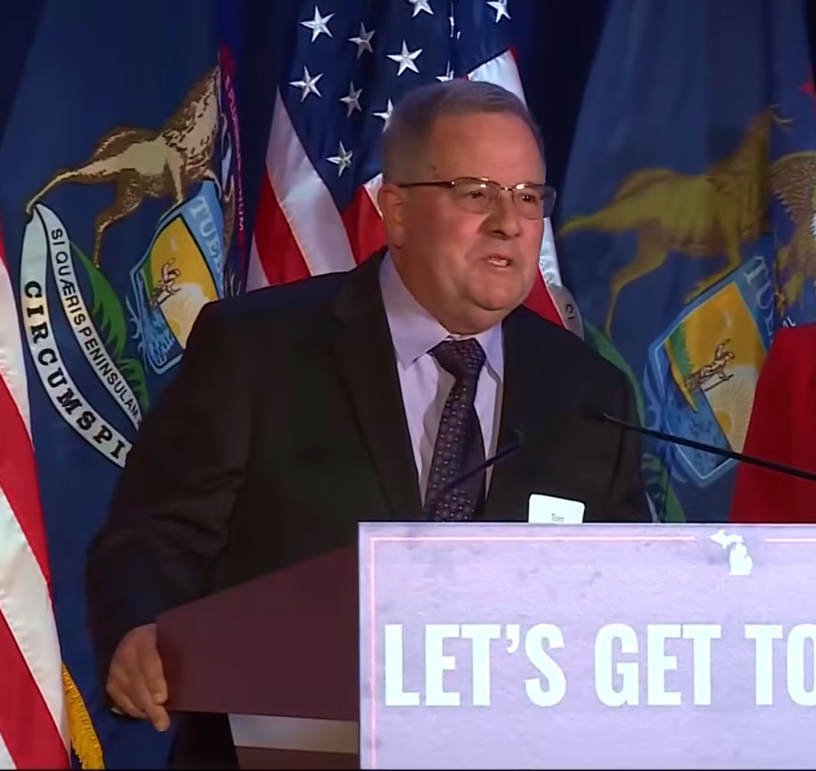 Thomas J. Smith speaking at a podium backed by American and Michigan flags